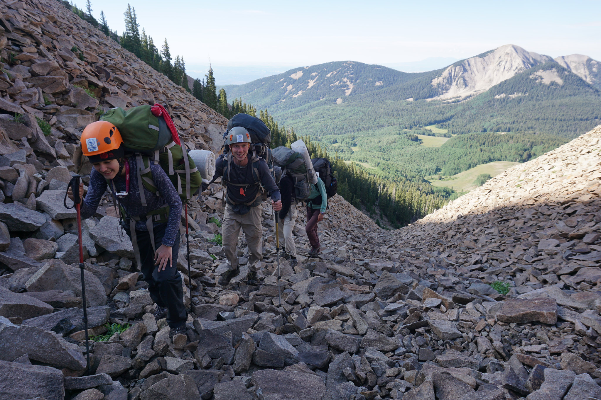 A group of students are hiking up a field of large, loose rock above treeline. They are wearing helmets and using trekking poles while carrying backpacks.